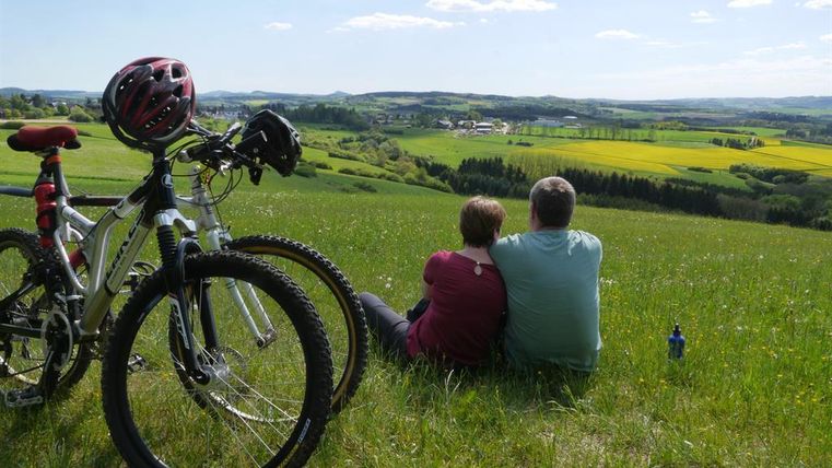 A couple is sitting on a meadow and enjoying the view of a green landscape. Next to them are two bicycles.