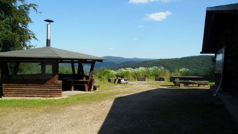 A generous view of a hilly landscape with a barbecue area and a wooden gazebo. The sky is clear and the surroundings are green.