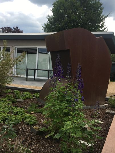 Une sculpture moderne en métal se trouve dans un jardin rempli de plantes. À l'arrière-plan, un bâtiment avec de grandes fenêtres est visible.