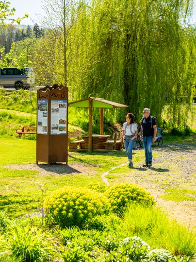 Ein Paar spaziert an einem Informationsschild und einem Pavillon in einem grünen Park vorbei.