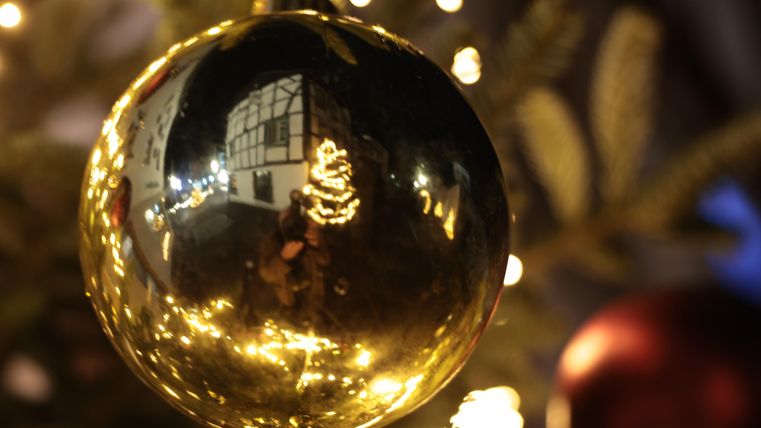 Een glanzende gouden kerstbal hangt aan een boom.
Op de achtergrond weerkaatst warm licht en een kerstdecoratie.