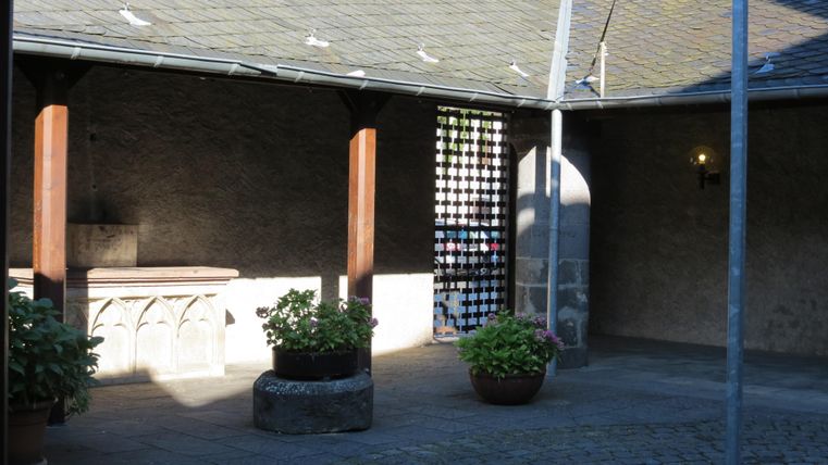 Ein ruhiger Innenhof mit steinernen Wänden und bepflanzten Kübeln. Die Sonne scheint, und das Licht schafft eine angenehme Atmosphäre.