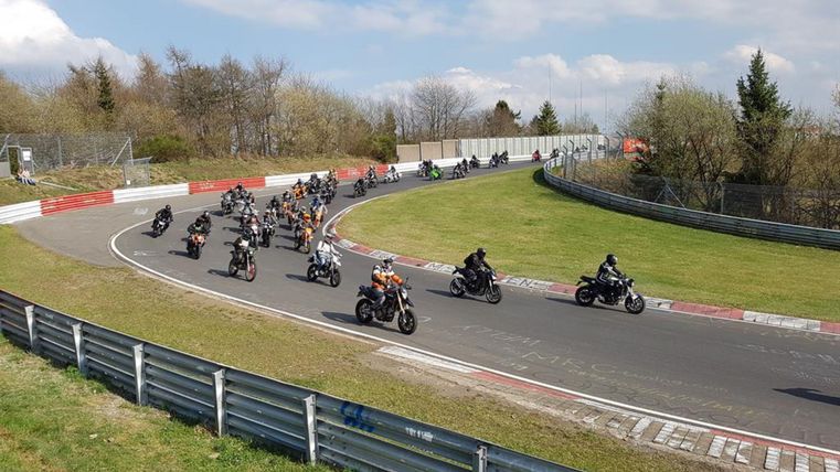 Eine Gruppe von Motorrädern fährt auf einer Rennstrecke durch eine Kurve. Die Strecke ist von Bäumen und Gras umgeben, unter einem klaren Himmel.