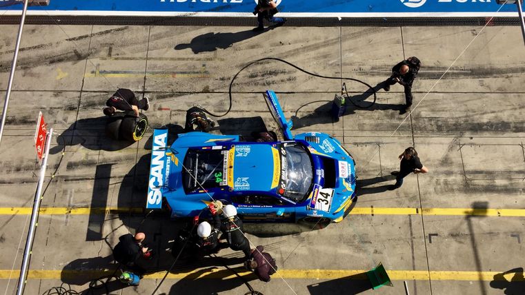 Ein blaue Rennwagen steht in der Boxenanlage, während ein Team von Mechanikern schnelle Wartungsarbeiten durchführt. Die Szene zeigt intensive Aktivität und Konzentration.