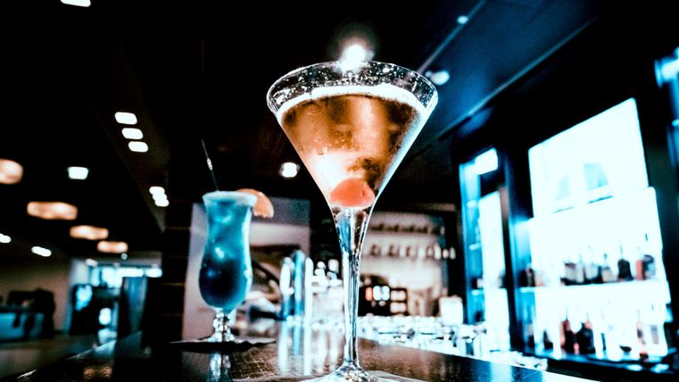An elegant cocktail in a martini glass, decorated with a cherry. In the background, more drinks can be seen on a bar.