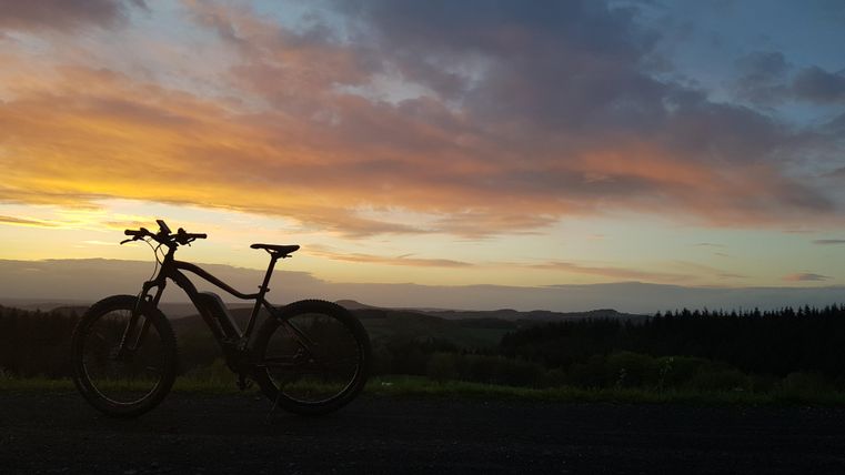 Ein Mountainbike steht im Vordergrund vor einem wunderschönen Sonnenuntergang. Die Wolken sind in warmen Farben gefärbt und die Landschaft ist im Hintergrund sichtbar.