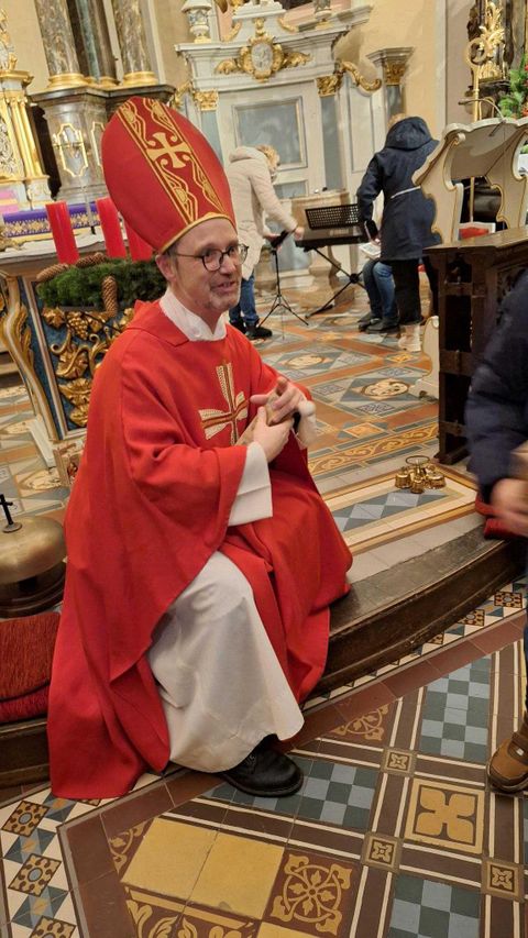 Een man in een rode mantel en mijter zit in een kerk. Hij glimlacht en lijkt met bezoekers te interageren.