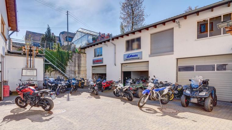 Eine Motorradausstellung mit vielen verschiedenen Bikes steht in einem Hof. Es gibt mehrere Gebäude im Hintergrund und grünes Laub.