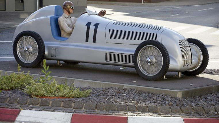 Ein silbernes Rennauto steht auf einem Sockel. Eine Figur sitzt im Auto und zeigt nach vorne.