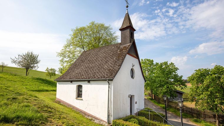 Eine kleine Kirche auf einem sanften Hügel, umgeben von grünen Bäumen. Der Himmel ist blau mit einigen Wolken.