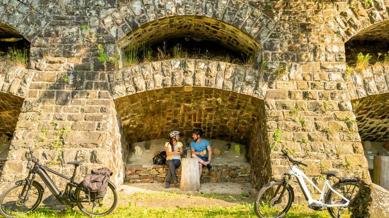 Ein Paar sitzt unter einem alten Bogen aus Stein und genießt die Aussicht. Neben ihnen stehen zwei Fahrräder auf dem Gras.