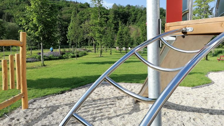 Ein Spielplatz mit einer Rutsche und weichem Sand. Im Hintergrund sind Bäume und eine grüne Wiese zu sehen.