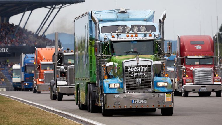 Eine Kolonne von bunten Lastwagen fährt auf einer Rennstrecke. Im Hintergrund sind Zuschauertribünen zu sehen.