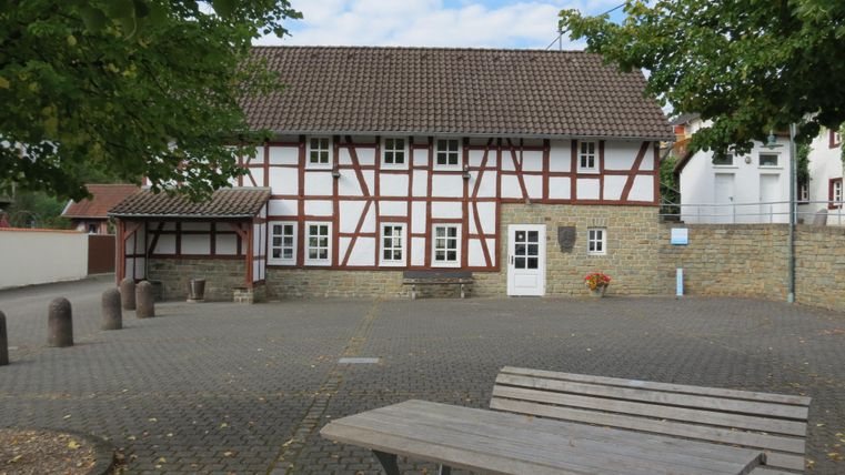 Ein historisches Fachwerkhaus mit einer gepflasterten Fläche davor. Im Hintergrund sind einige Bäume und ein bewölkter Himmel zu sehen.