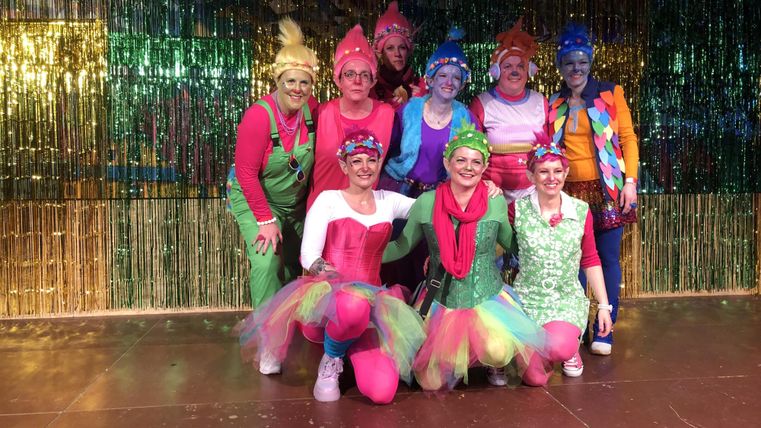 Een groep vrouwen in kleurrijke kostuums staat op het podium. Ze glimlachen vrolijk voor een glinsterende achtergrond.