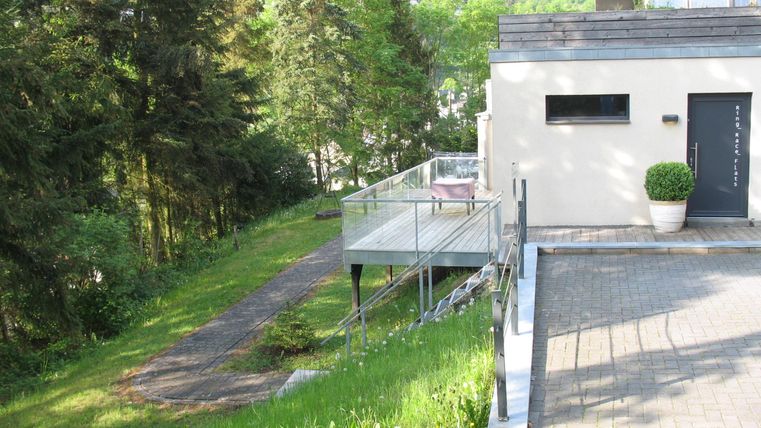 Ein modernes Haus mit einer Terrasse und einem schönen, grünen Garten. Im Hintergrund sind Bäume und Hügel zu sehen.