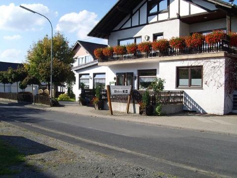 Ein gemütliches Haus mit einem Balkon, der mit Blumen dekoriert ist. Die Straße führt an dem Haus vorbei und es ist sonnig.