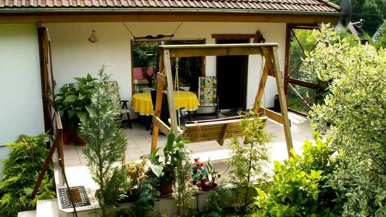 A cozy terrace with a swing and many flowers. The view shows an inviting residential house surrounded by greenery.