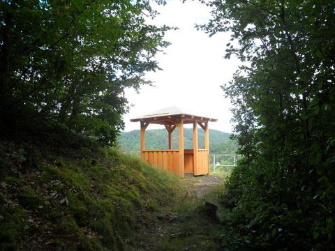 Ein Holzpavillon steht am Ende eines schmalen Weges, umgeben von Bäumen. Im Hintergrund sind sanfte Hügel sichtbar.