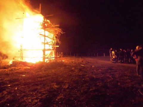 Ein großes Feuer brennt an einem Gerüst in der Nacht. Eine Gruppe von Personen beobachtet das Geschehen in sicherer Entfernung.