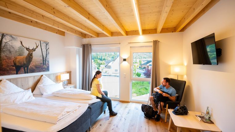 A modern room with a wooden ceiling, two beds, and a large window. Two people are sitting relaxed in the room and are having a conversation.