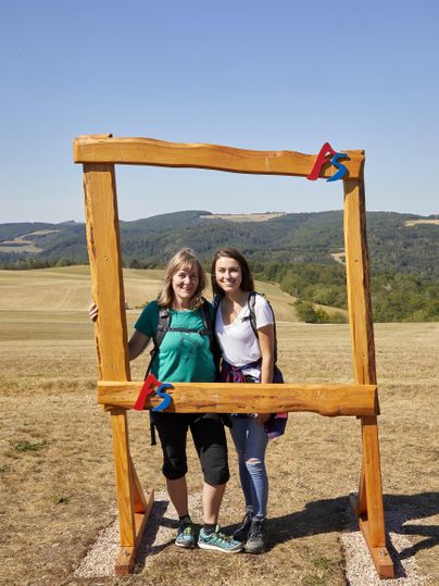 Zwei Frauen stehen in einem Holzrahmen mit einer Landschaft im Hintergrund. Es ist ein sonniger Tag mit klarem Himmel.