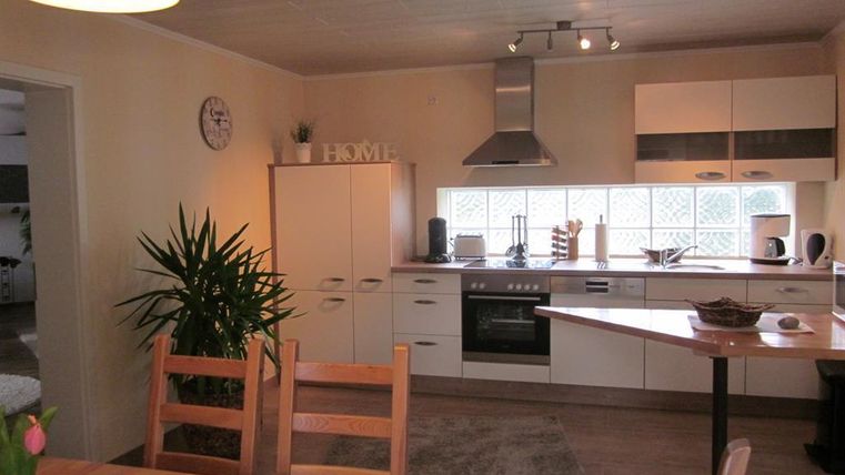 Een heldere, moderne keuken met eenvoudige meubels en een groot raam. Op de voorgrond staat een houten tafel met stoelen en een plant.