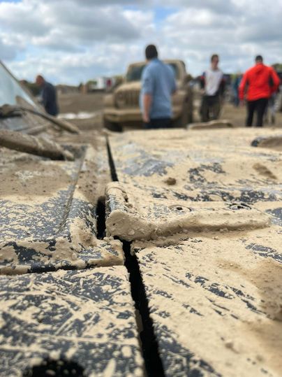 Ein verschmutztes Fahrzeug im Vordergrund, umgeben von Menschen auf einem schmuddeligen Gelände. Der Himmel ist bewölkt und es herrscht eine lebendige Atmosphäre.