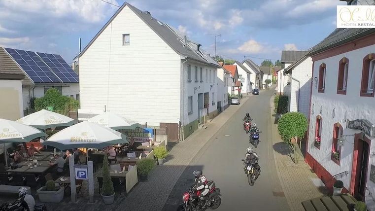 A quiet street in a small village with cafes and motorcycles. The houses are bright and there is a view of the sky.