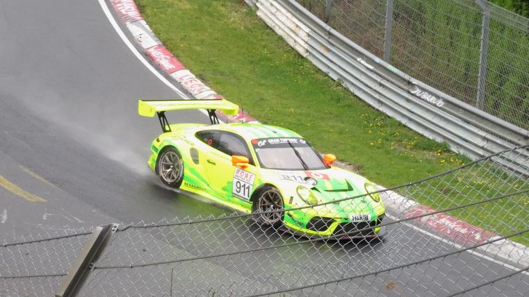 Une voiture de course roule sur un circuit sinueux. Elle a une peinture verte frappante et des éclaboussures d'eau sont visibles.