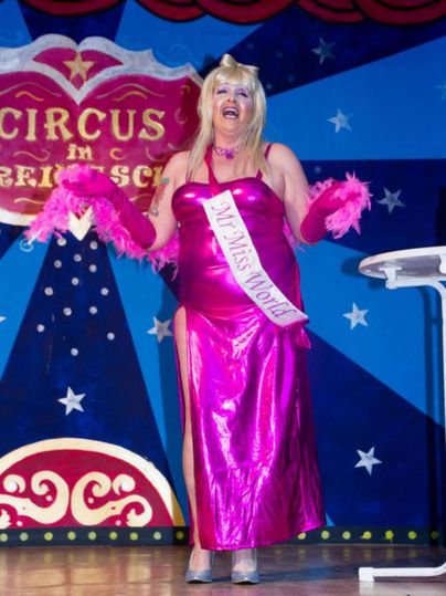 Een persoon in een opvallende, roze jurk staat op het podium en glimlacht. Ze draagt een bord met de tekst "Miss World" en roze accessoires.