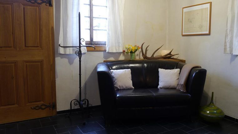 A cozy living room with a dark brown sofa and two cushions. Curtains hang on the wall, and there is a decorative hanging as well as a flower arrangement on the table.