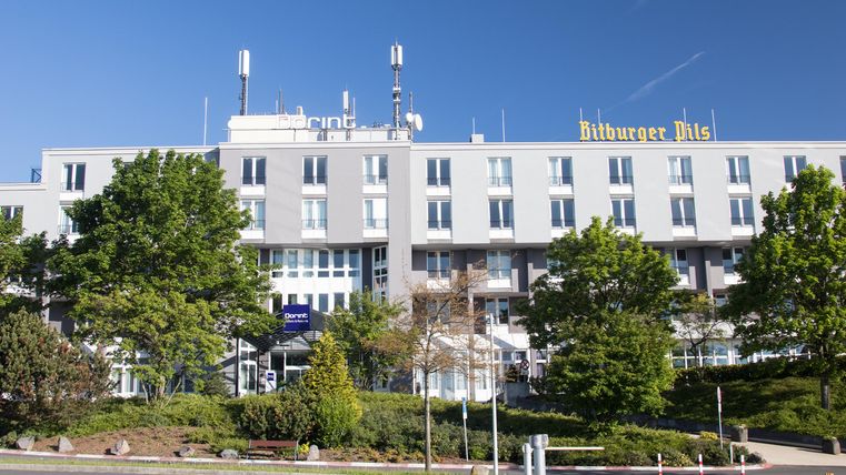 Een modern hotelgebouw met veel ramen en een heldere blauwe lucht. Omgeven door groene bomen en goed onderhouden terrein.