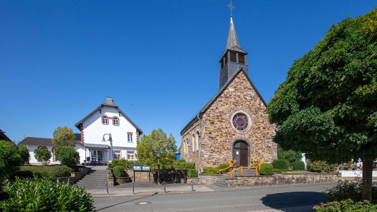 Een schilderachtige straat met een kerk en een wit gebouw. De omgeving is groen en zonnig, met een heldere blauwe lucht.