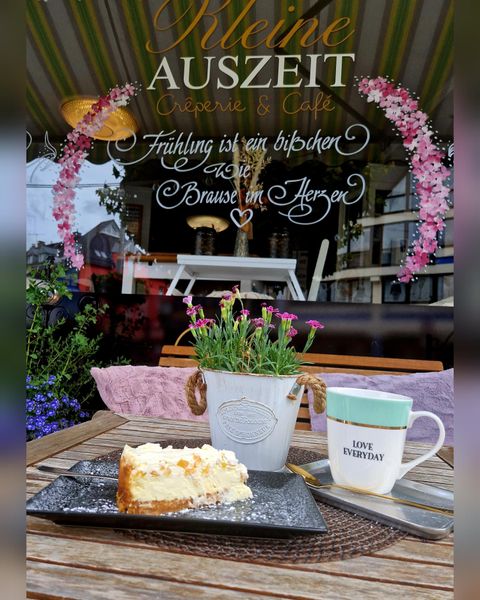 Ein schöner Tisch mit einem Stück Käsekuchen und einer Tasse Kaffee. Im Hintergrund ist ein Fenster mit floralen Dekorationen und einem einladenden Schriftzug.