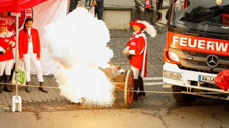 Eine Feuerwehrveranstaltung mit einem Festumzug. Eine Kanone stößt Rauch aus, während die Teilnehmer in roten Kostümen stehen.