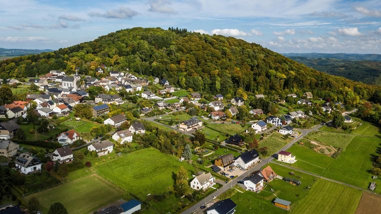 Luftbild des Dorfes Aremberg mit grünen Wiesen und bewaldetem Hügel im Hintergrund.