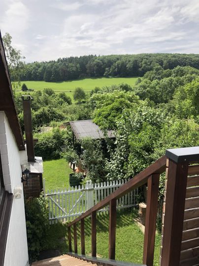 A peaceful garden landscape with plenty of greenery and a small shed. In the background, gentle hills and forests can be seen.