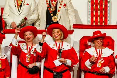 Drei Frauen in roten Kostümen und Hüten feiern fröhlich. Sie halten Gläser in der Hand und lächeln in die Kamera.