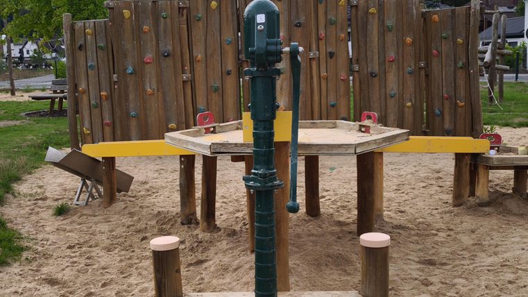 Ein Spielplatz mit einer Kletterwand und einem Wasserspielgerät steht in einer sandigen Umgebung. Im Hintergrund sind grüne Bäume und eine Wohngegend sichtbar.