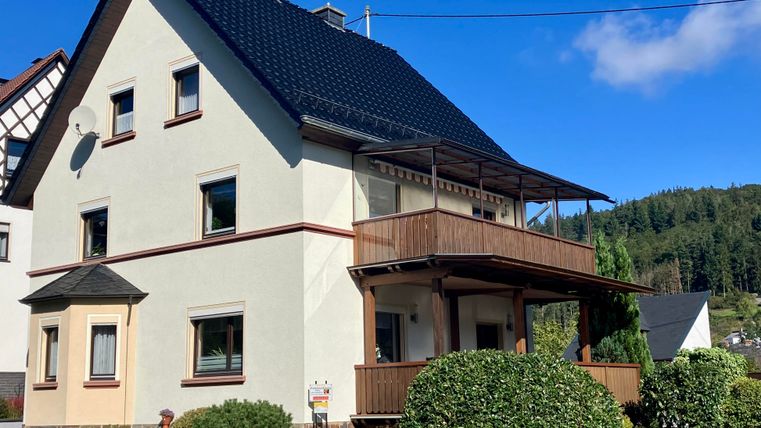 Une maison moderne avec un toit noir et un balcon. Entourée d'un jardin entretenu et d'un ciel bleu clair.