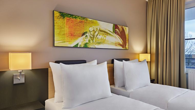 A modern hotel room with two single beds and soft color tones. A colorful painting hangs on the wall, and the room is bright and inviting.