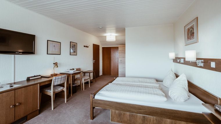 Een eenvoudig hotelkamer met twee eenpersoonsbedden, een televisie en een tafel. De muren zijn licht en de inrichting is modern en functioneel.