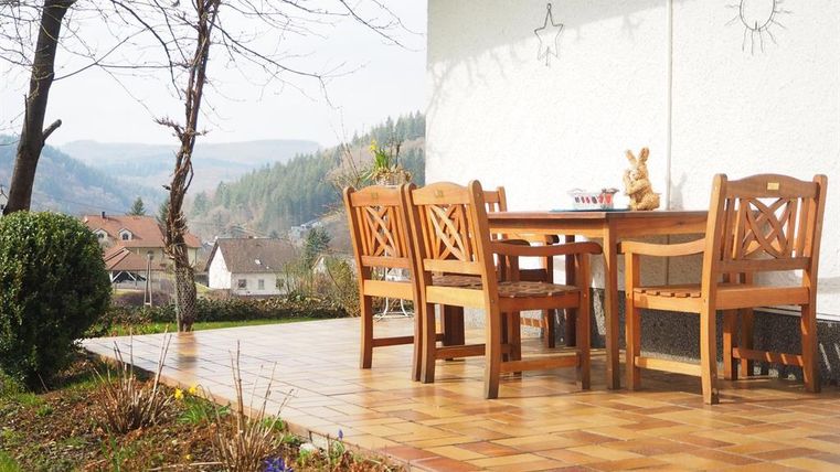 Een gezellige buitenruimte met een houten tafel en stoelen. Op de achtergrond zijn heuvels en huizen te zien.