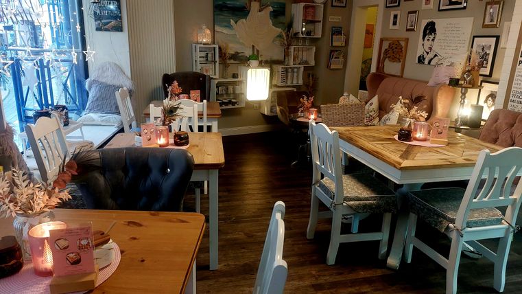 Un café confortable avec des tables et des chaises en bois clair. Les murs sont décorés avec des tableaux, et quelques bougies ajoutent une atmosphère accueillante.