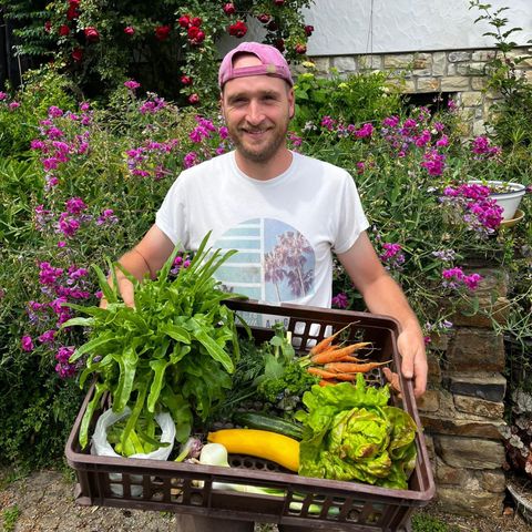 A smiling man holds a basket of fresh vegetables and herbs. Colorful flowers are blooming in the background.