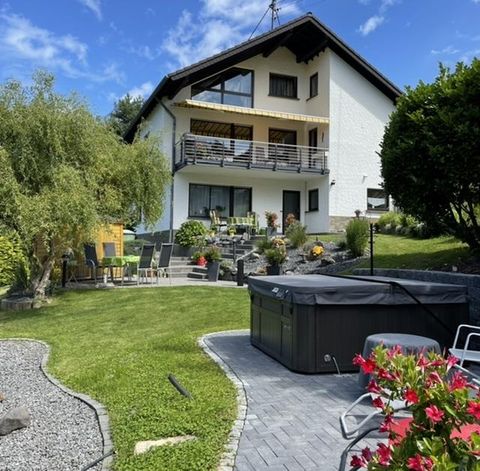 A beautiful house with a well-maintained garden and a terrace. In the foreground, there is a hot tub and colorful flower beds.