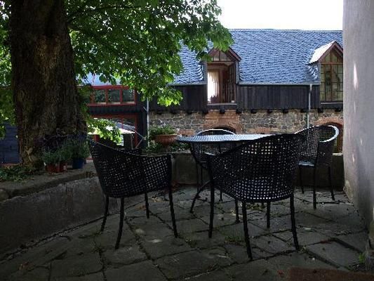 Ein gemütlicher Außenbereich mit schwarzen Korbsesseln und einem runden Tisch. Im Hintergrund sind Gebäude mit Holzverkleidung und einem grünen Baum zu sehen.