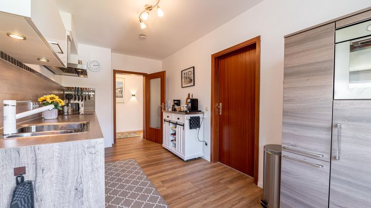 Een moderne keuken met houten vloer en witte meubels. Er is een koelkast, een fornuis en veel daglicht.