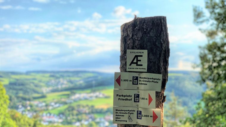 Een houten wegwijzer staat in een mooie landschap. Op de achtergrond zijn zachte heuvels en een blauwe hemel te zien.